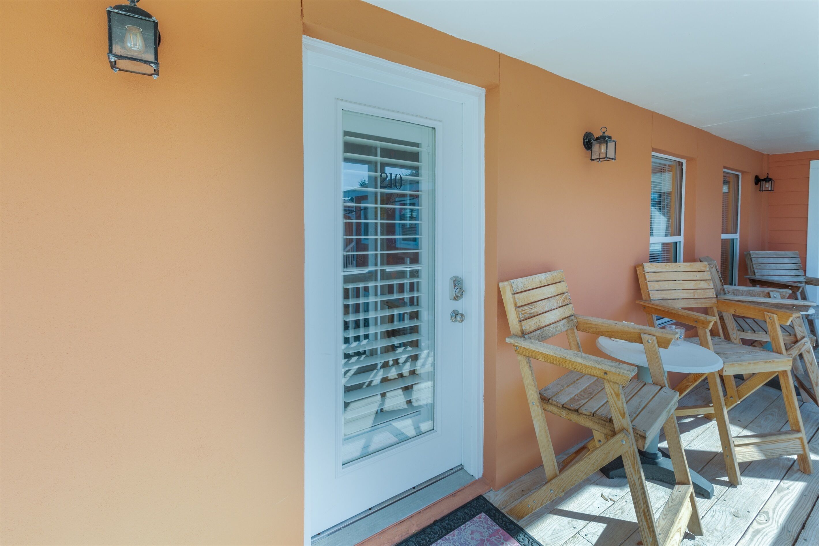 A wooden porch with chairs and a table, a white door with a window, and a wall-mounted light fixture.