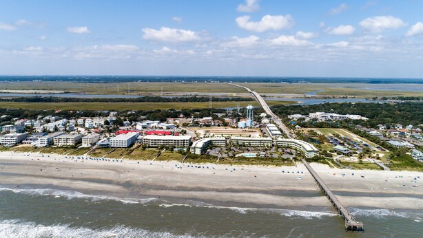 An aerial view of a coastal area with a beach, a pier, and a hotel complex with multiple buildings and a swimming pool.