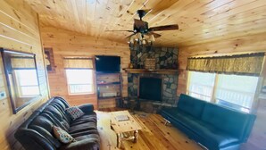 L’intérieur chaleureux d’un chalet avec une cheminée en pierre, un ventilateur de plafond et un plancher en bois.