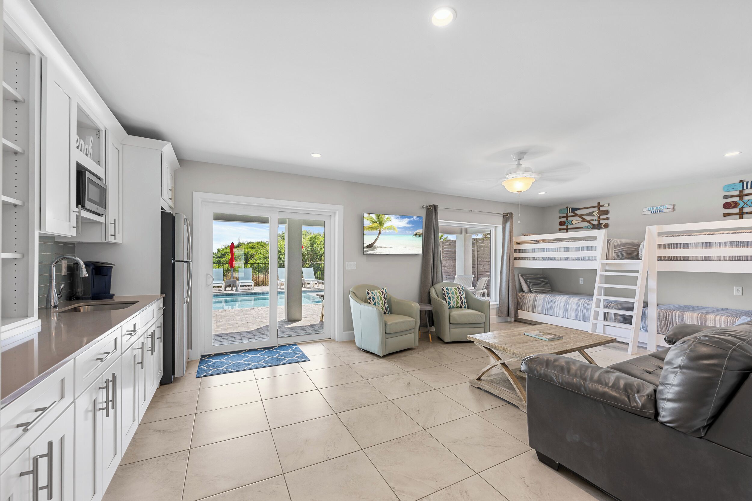 A modern kitchen with white cabinets, a built-in microwave, and a sink. There is a pool visible through the glass doors, and a living area with a sofa and a coffee table.