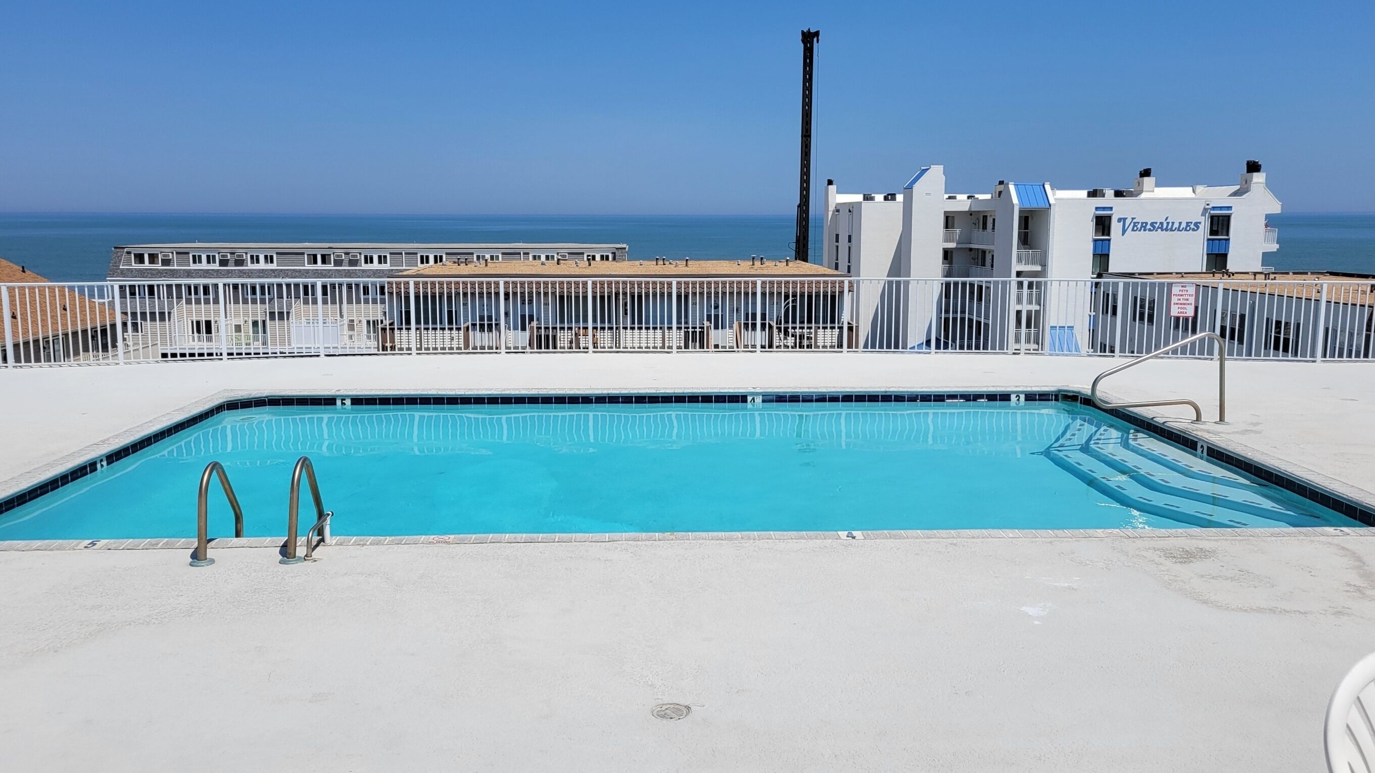 A swimming pool with a metal ladder, a fenced area, and a building with a sign reading "VERSAILLES" in the background.