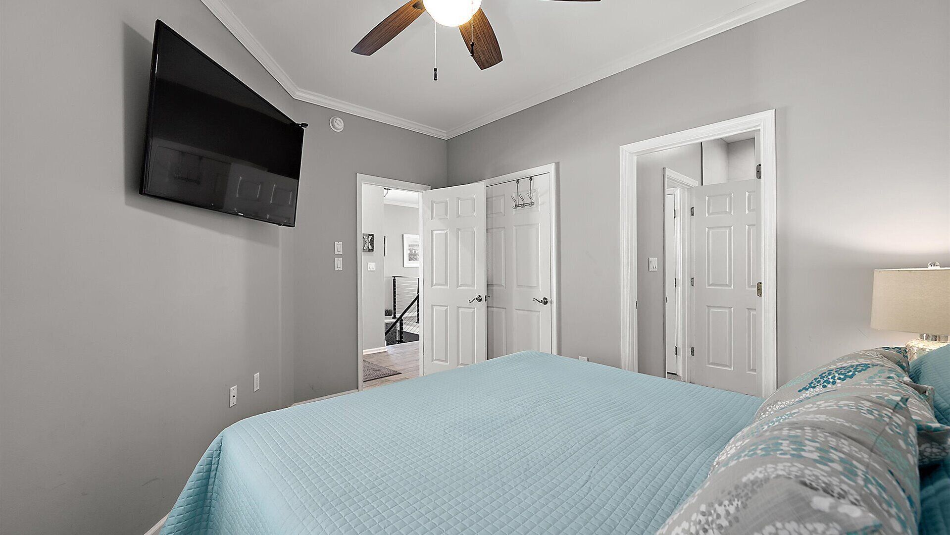 A bedroom with a bed, a ceiling fan, a TV mounted on the wall, and two doors leading to other rooms.