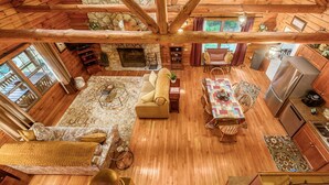 A spacious cabin interior with a stone fireplace, wooden floors, a dining area, and a kitchen.