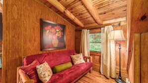 A wooden cabin interior with a red sofa, a painting of flowers, a floor lamp, and a window with curtains.