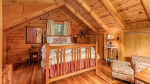 A cozy wooden cabin bedroom with a bed, a chair, a nightstand, and a window with curtains.