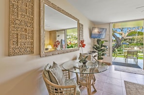 A dining area with a glass table and wicker chairs, a large ornate mirror, and a view of the outdoor patio with a tree and seating. - Beautiful 1BR Poolside Condo E141 w/AC, Sleeps 4, Near Napili Bay & Turtle Cove (Lahaina)