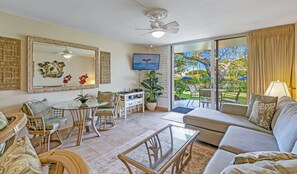 A spacious living area with a glass coffee table, wicker chairs, a sofa, and a dining table. There is a television mounted on the wall, a ceiling fan, and a large window with curtains. - Beautiful 1BR Poolside Condo E141 w/AC, Sleeps 4, Near Napili Bay & Turtle Cove (Lahaina)