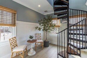 Une pièce dotée d’un escalier en colimaçon, d’un arbre en pot, de deux fauteuils en osier, d’une petite table et d’une fenêtre avec des stores.