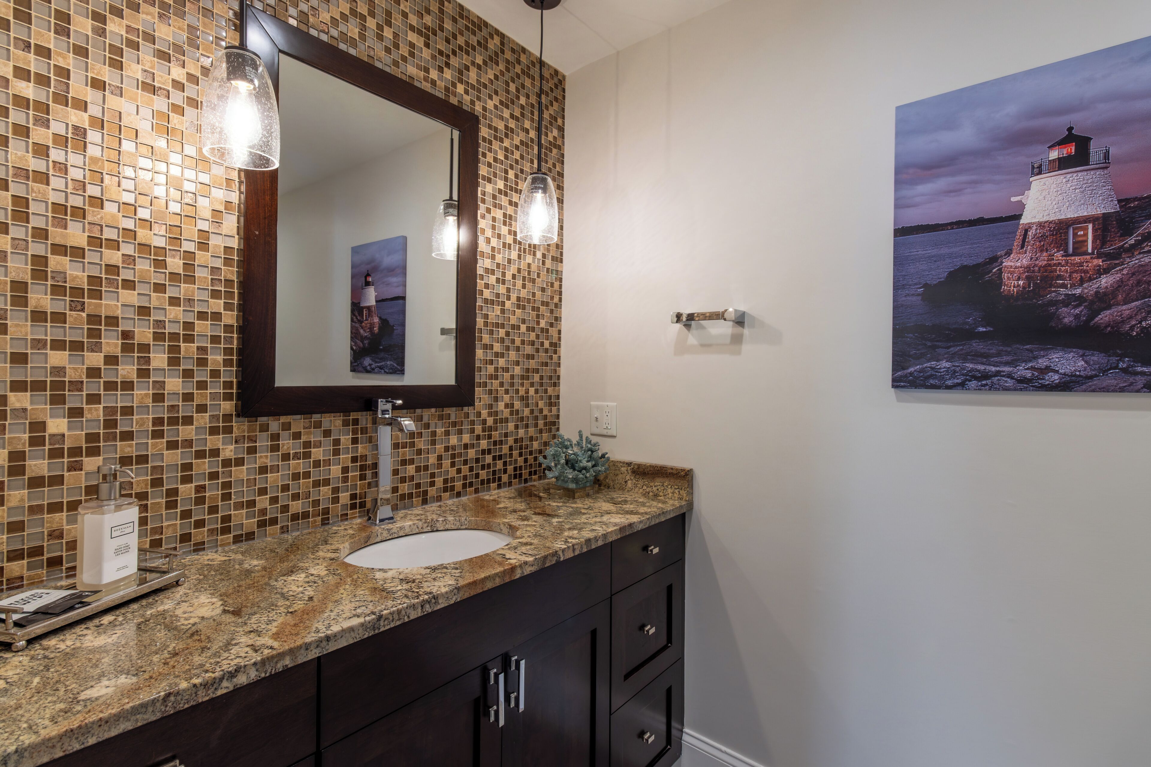 Ein Badezimmer mit Mosaikfliesen an der Rückwand, einer Granit-Arbeitsplatte, einem weißen Waschbecken, dunklen Holzschränken und einem gerahmten Foto eines Leuchtturms.