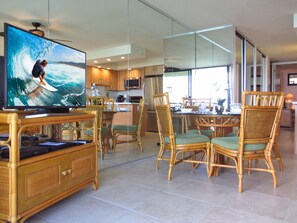 A modern kitchen with a dining area, featuring a large flat-screen TV, wicker furniture, and a view of the ocean.