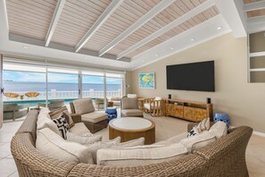 A spacious living area with a large window overlooking a pool and the sea, furnished with wicker sofas and a flat-screen TV mounted on the wall. - Private Cable Beach villa with pool, oceanfront terrace & guest cottage (Cable Beach, Nassau)