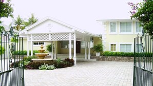 A gated entrance with a covered porch, a fountain, and a paved driveway leading to a building with a white facade and a yellow accent wall. - Luxury Beach--front house, perfect location! Cable Beach BEAUT! Walk to Baha Mar (Cable Beach, Nassau)