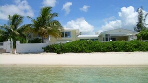 A beachfront property with a white sandy beach, clear turquoise water, and a two-story house with a covered porch. - Luxury Beach--front house, perfect location! Cable Beach BEAUT! Walk to Baha Mar (Cable Beach, Nassau)