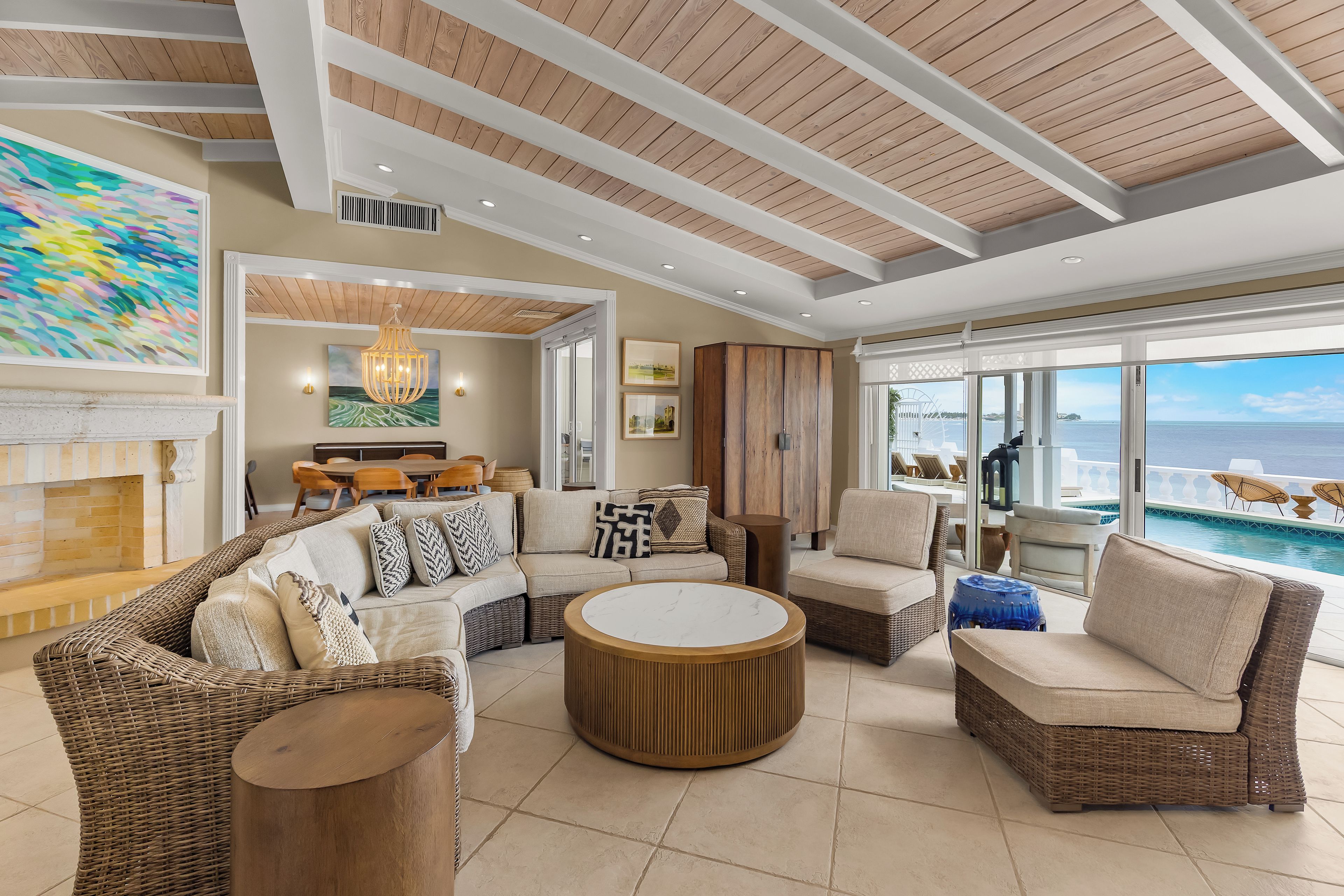 A spacious living area with a large sofa, round coffee table, and wicker furniture. There is a dining area with wooden chairs and a table, and a view of the ocean through large windows.