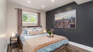 Ein Schlafzimmer mit einem Bett, einem Nachttisch, einem Fenster mit Vorhängen, einem Stadtbildgemälde an der Wand und einem Holzboden.