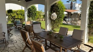 Een terras buiten met een houten tafel en stoelen, omringd door witte zuilen en overdekt met een dak.
