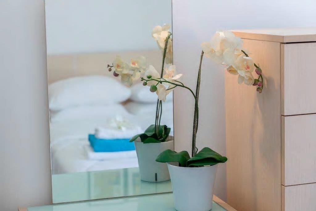A mirror reflecting a bed with white pillows and a blue object on it, and two white orchids in white pots on a glass surface.