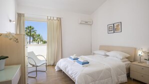 Een hotelkamer met een bed, nachtkastje, stoel en uitzicht op palmbomen door het raam.