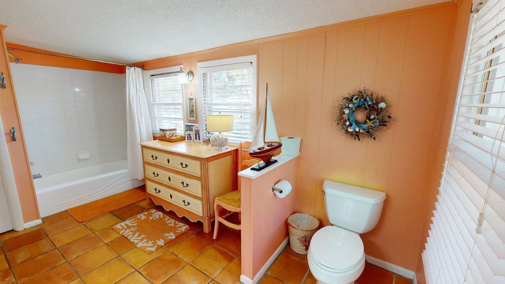 A bathroom with a bathtub, a toilet, a vanity with drawers, a small table with a lamp, and a decorative wreath on the wall.