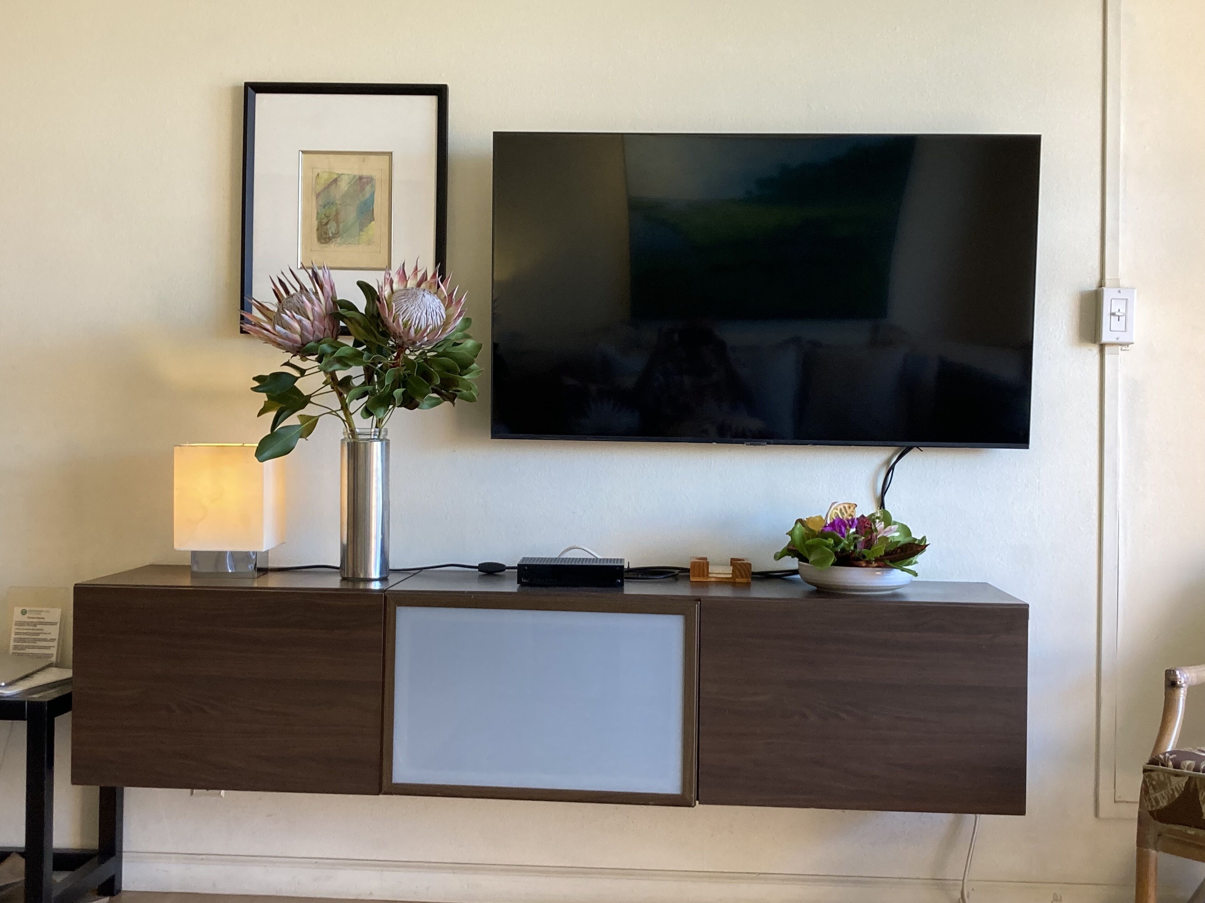 A modern living room with a flat-screen TV mounted on the wall, a wooden cabinet with a glass front, and a vase with flowers on the cabinet.
