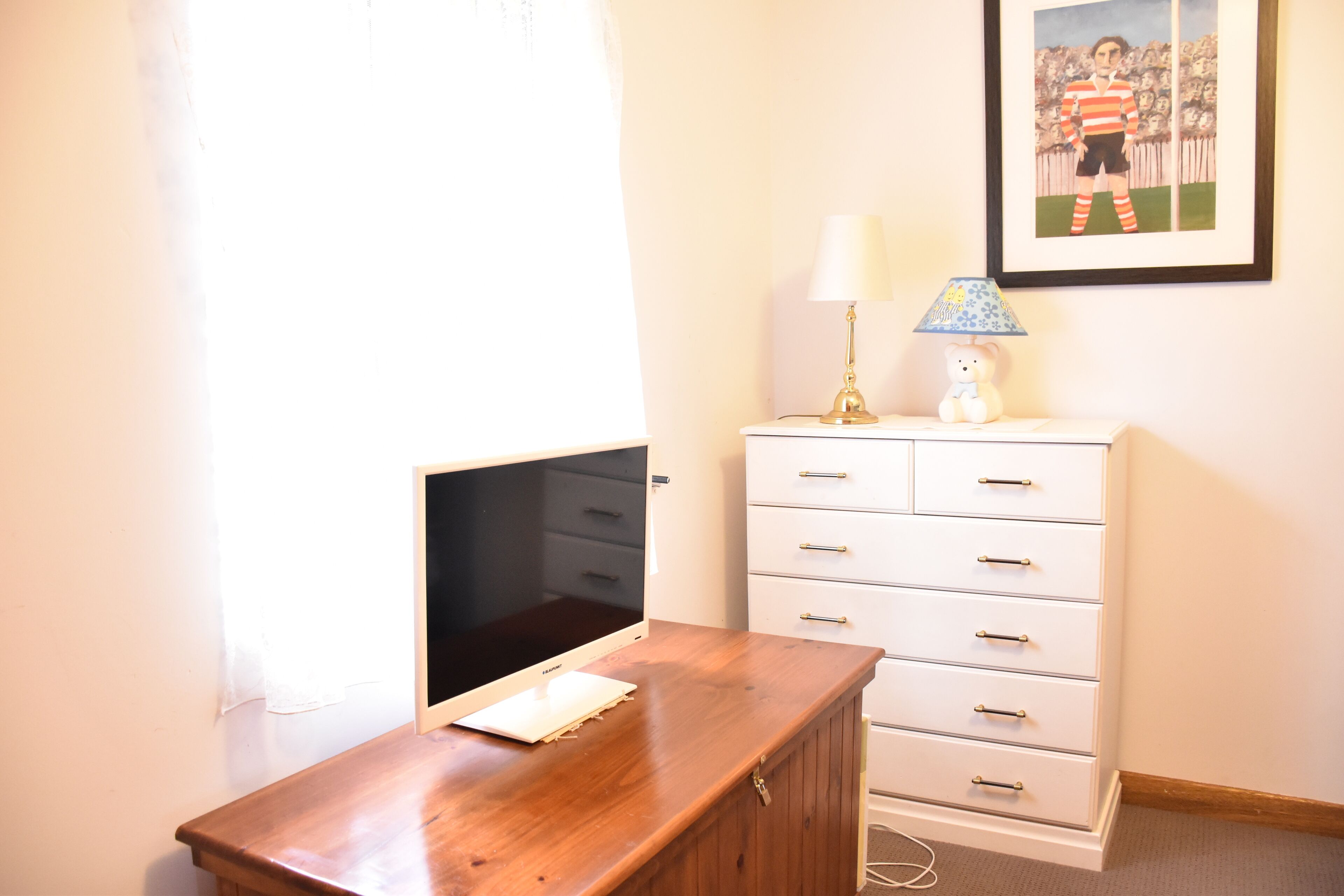 Une pièce comprenant un bureau en bois, un téléviseur, une commode blanche avec des tiroirs, une lampe et un tableau encadré accroché au mur.