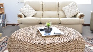 A living room with a beige sofa, a round woven ottoman, a small potted plant, a remote control, and a magazine.