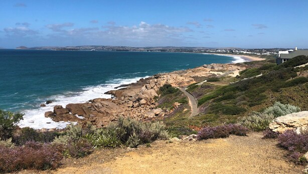 A coastal landscape with rocky terrain, a dirt path, and a view of the ocean and distant buildings.