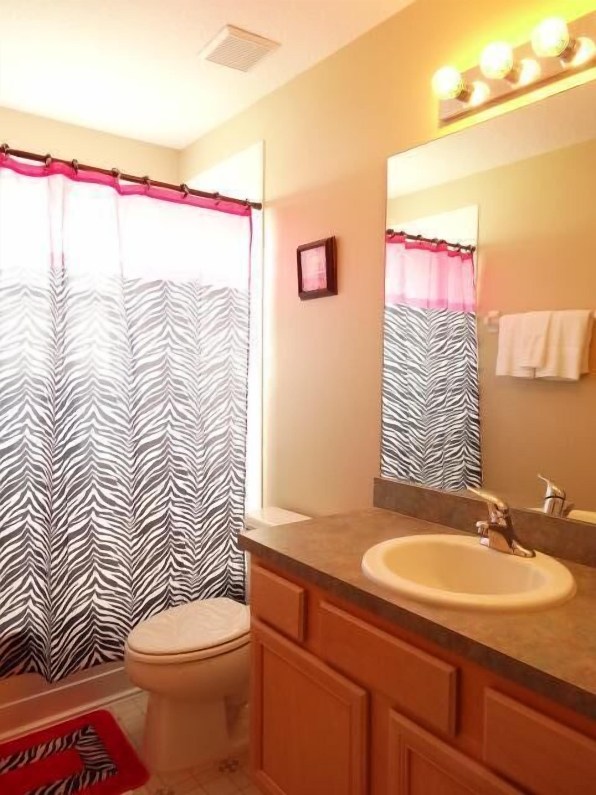 Un baño con cortina de ducha a rayas de cebra, un inodoro blanco, un lavabo beige con un espejo encima y una alfombra de baño roja.