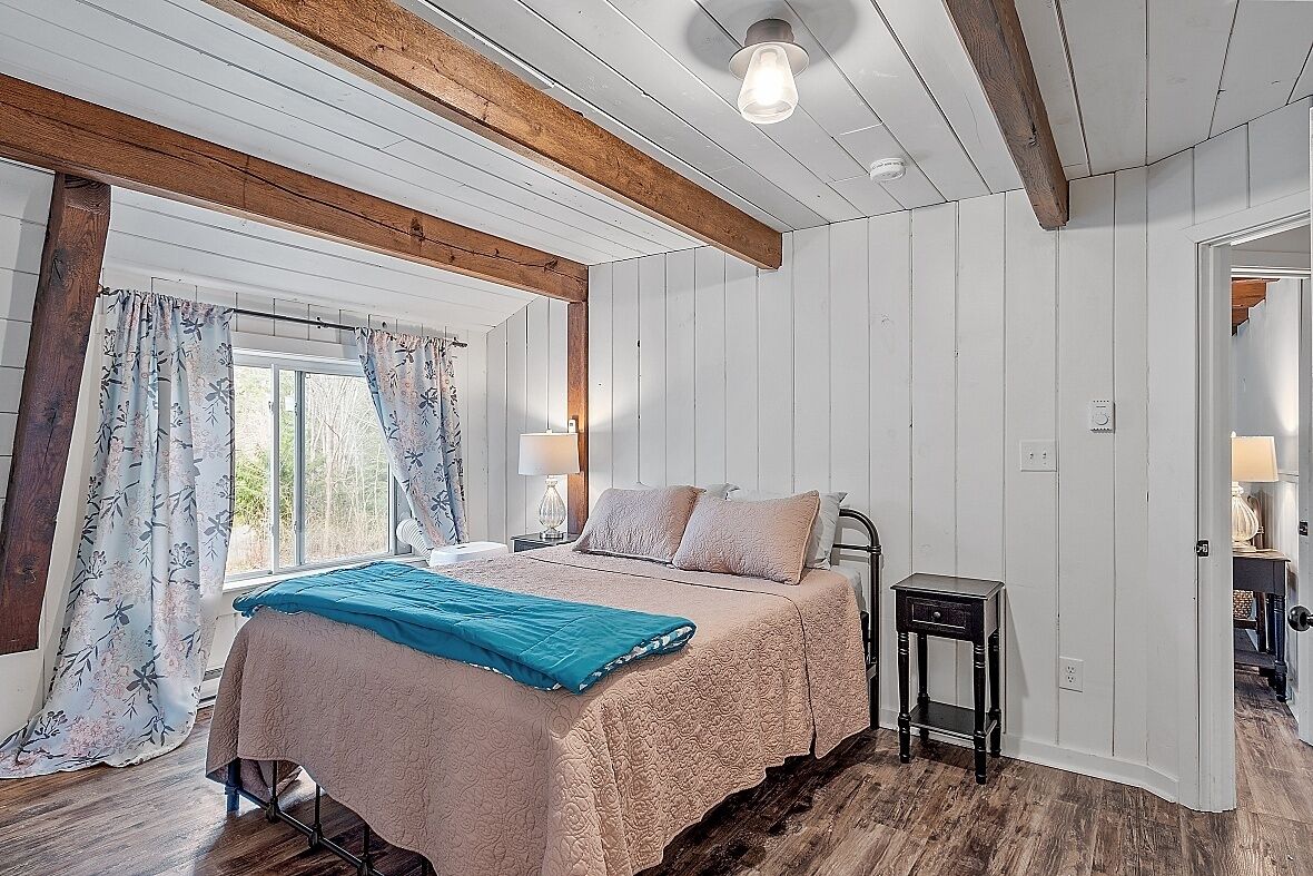 Une chambre à coucher avec un lit, des poutres en bois, une fenêtre avec des rideaux, une table de chevet et une commode.