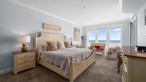 A bedroom with a large bed, bedside tables, a desk, and a view of the beach through large windows.