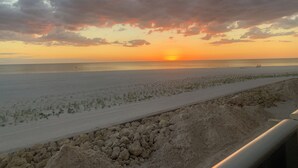 En strand med en solnedgång i bakgrunden, en sandstig och ett stenigt område i förgrunden.