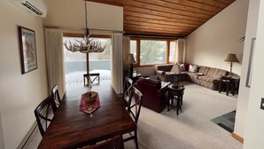 A dining area with a wooden table and chairs, a chandelier, and a view of the outdoors through large windows.