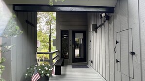 A covered porch with a wooden floor, a bench, and a planter with flowers and an American flag.