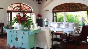 A spacious room with a high ceiling, wooden beams, a white sofa set, a turquoise cabinet, and a large window with a view of greenery. - Elegant Villa in Private Hope Bay. Infinity Pool with Sea View. (Bequia)