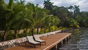 Una terraza de madera con sillones de descanso que se extiende sobre el agua, rodeada de exuberante vegetación y palmeras.