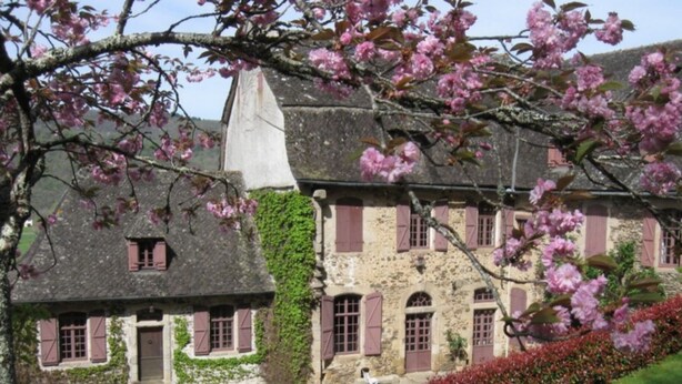 Un edificio de piedra con contraventanas rosas y techo de paja, rodeado de un jardín cuidado y un cielo despejado.