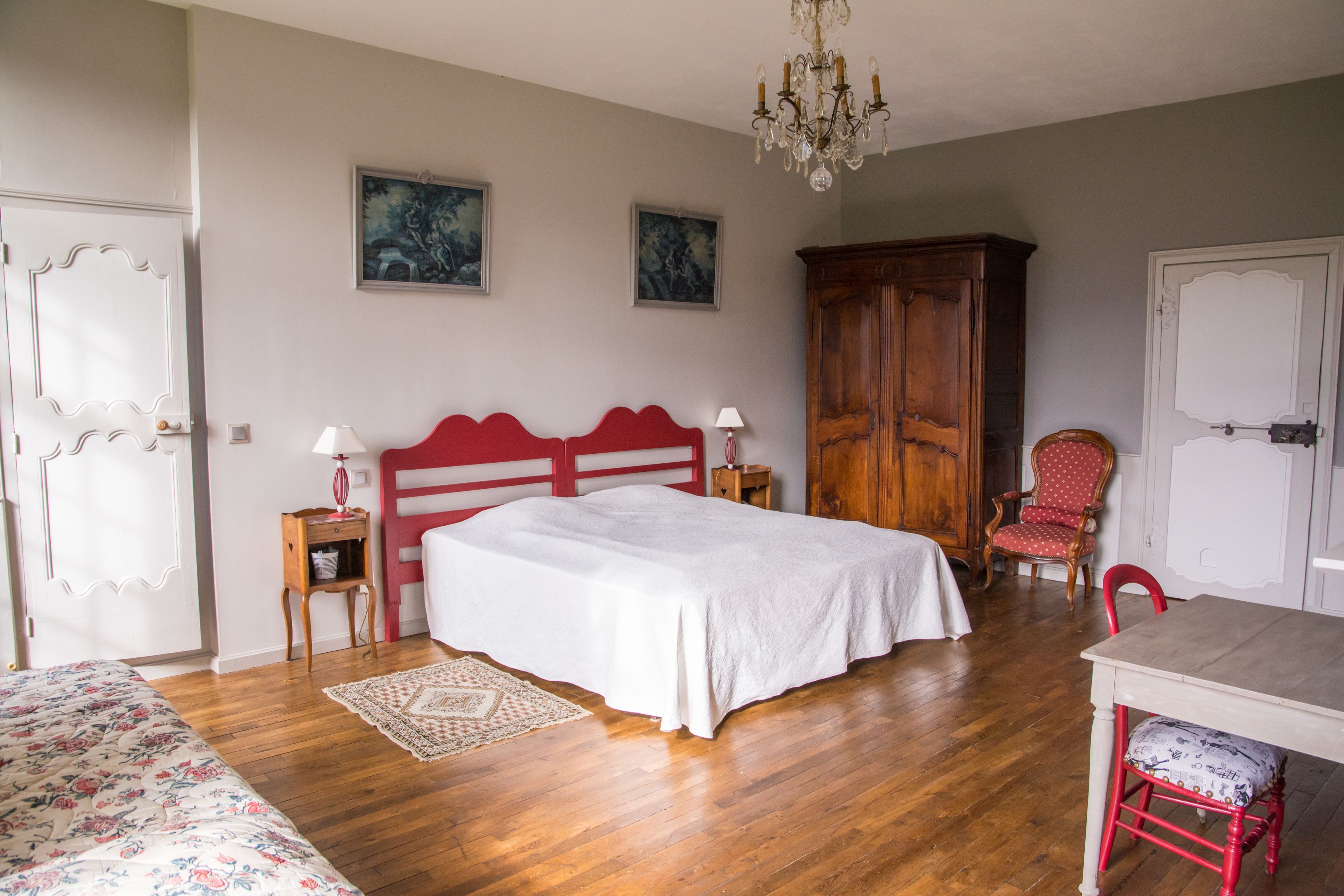 A bedroom with a wooden floor, a bed with a white bedspread, a wooden wardrobe, a red chair, and a chandelier.