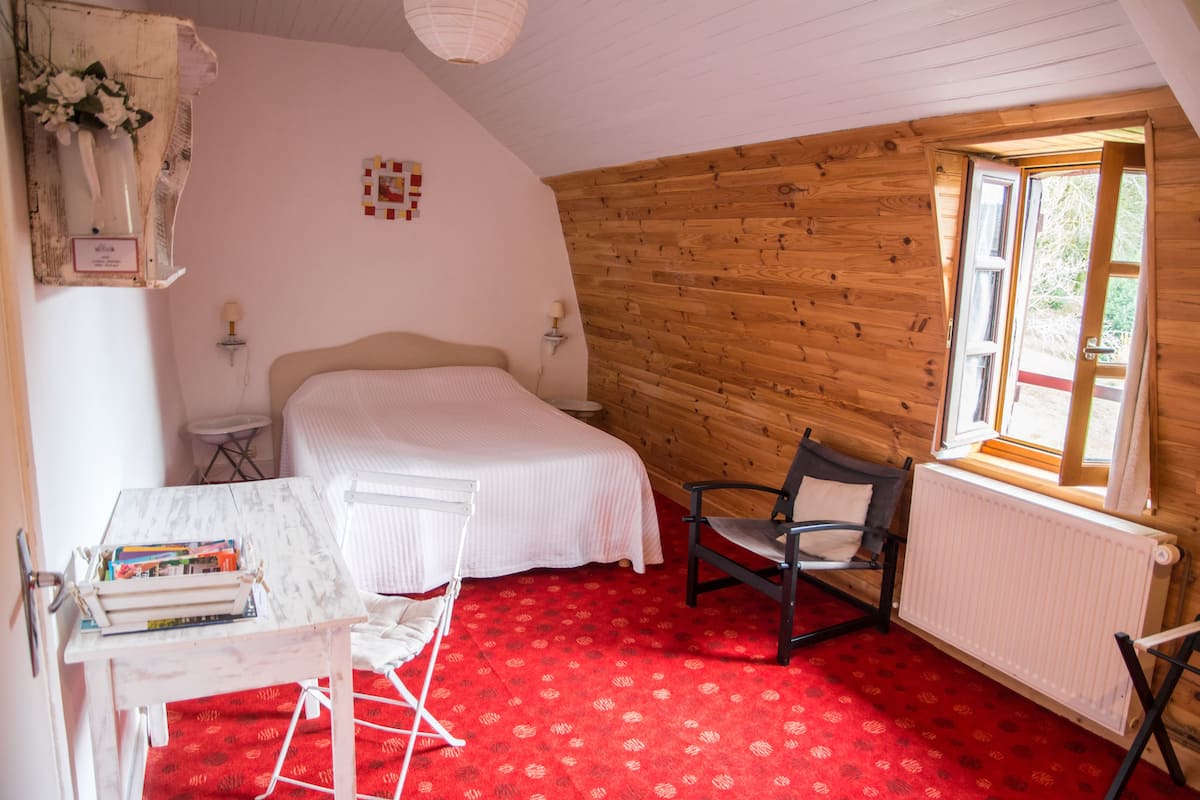 Ein gemütliches Schlafzimmer mit Holzbalkendecke, einem roten Teppich, einem Bett, einem Sessel, einem kleinen Tisch mit Büchern und einem Fenster mit Aussicht.