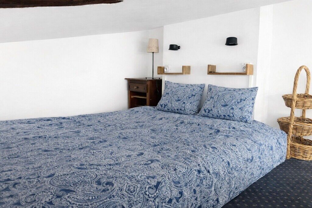 Un dormitorio con una cama grande, una mesita de noche, una lámpara, un estante en la pared con dos tazas y una cesta de mimbre.