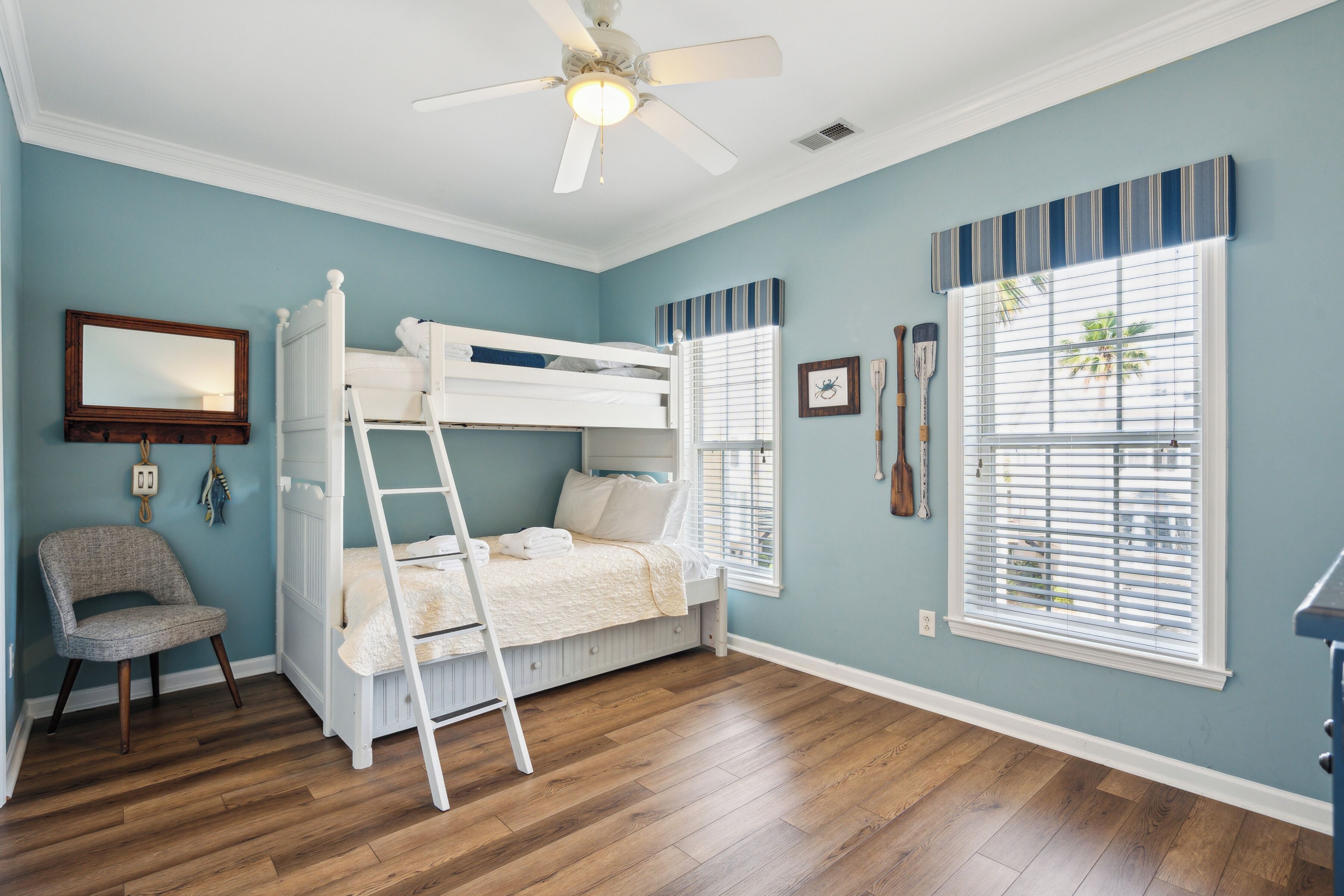 A bedroom with a bunk bed, a chair, a ladder, a ceiling fan, a window with blinds, and a wall-mounted mirror.