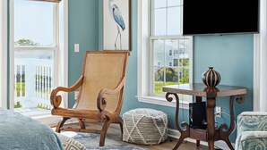 A bedroom with a large window, a wooden chair, a bedside table with a vase, and a framed picture on the wall. - Water's Edge 83 - Comfy Cool - Pool - River Views - Close to Everything - Beautifully Updated (Folly Beach)