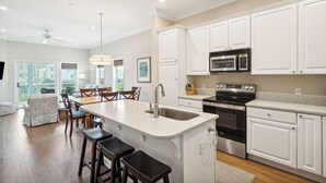 A modern kitchen with white cabinetry, a central island with stools, and a dining area with chairs and a table. - Water's Edge 83 - Comfy Cool - Pool - River Views - Close to Everything - Beautifully Updated (Folly Beach)