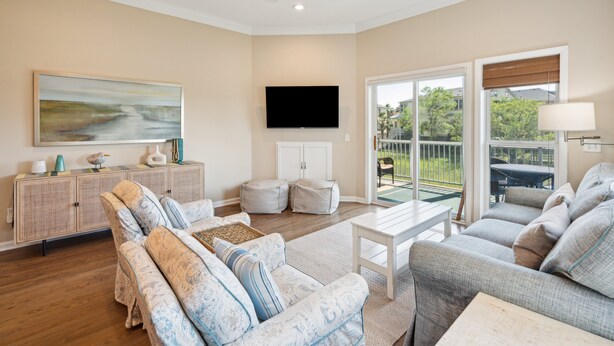 A spacious living room with a large flat-screen TV, a ceiling fan, and a view through glass doors to an outdoor area. - Water's Edge 83 - Comfy Cool - Pool - River Views - Close to Everything - Beautifully Updated (Folly Beach)