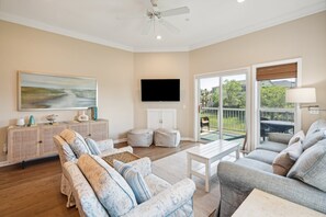 A spacious living room with a large flat-screen TV, a ceiling fan, and a view through glass doors to an outdoor area.