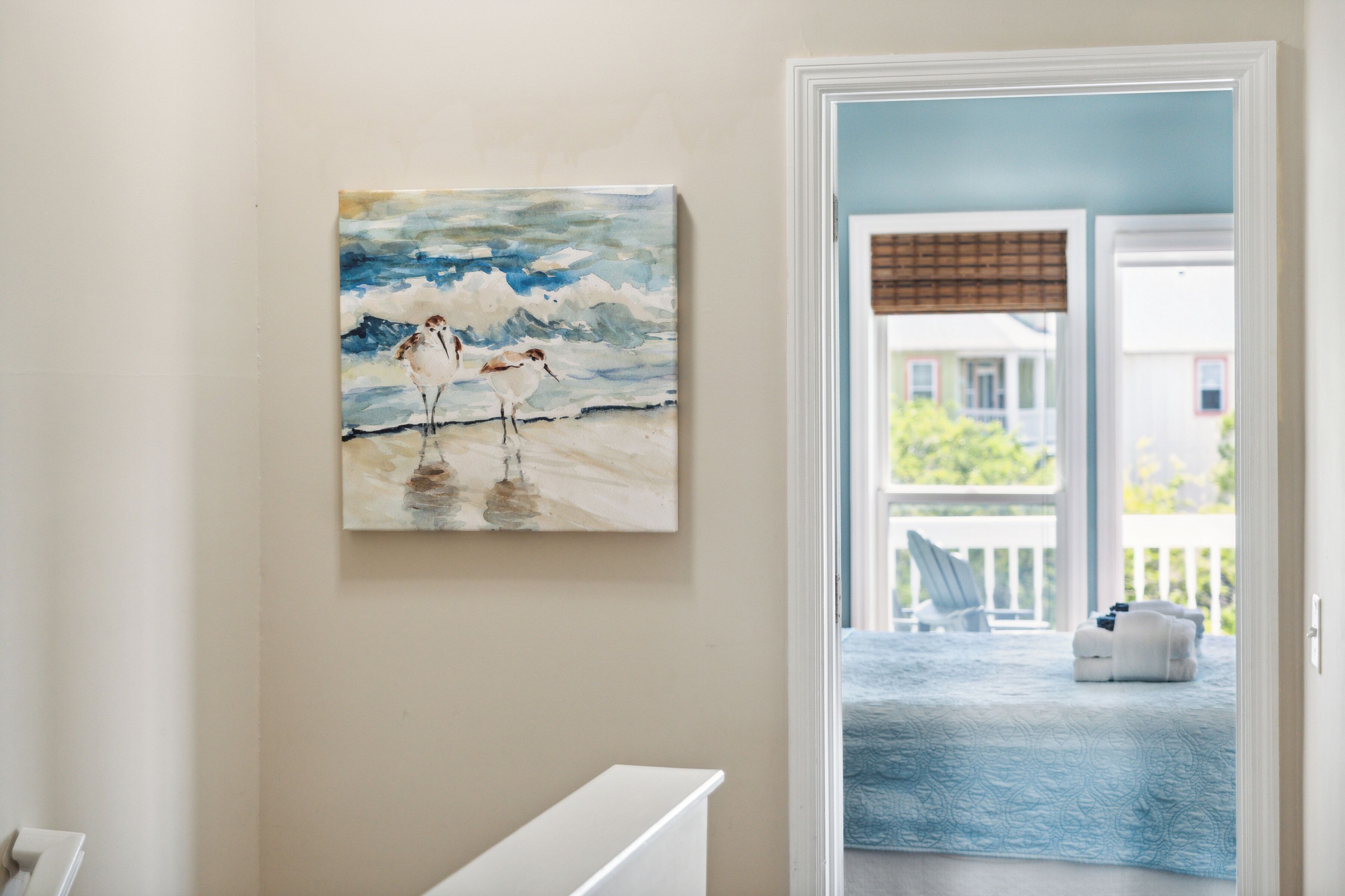 A room with a painting of birds on a wall, a white door leading to a balcony, and a blue patterned bedspread.