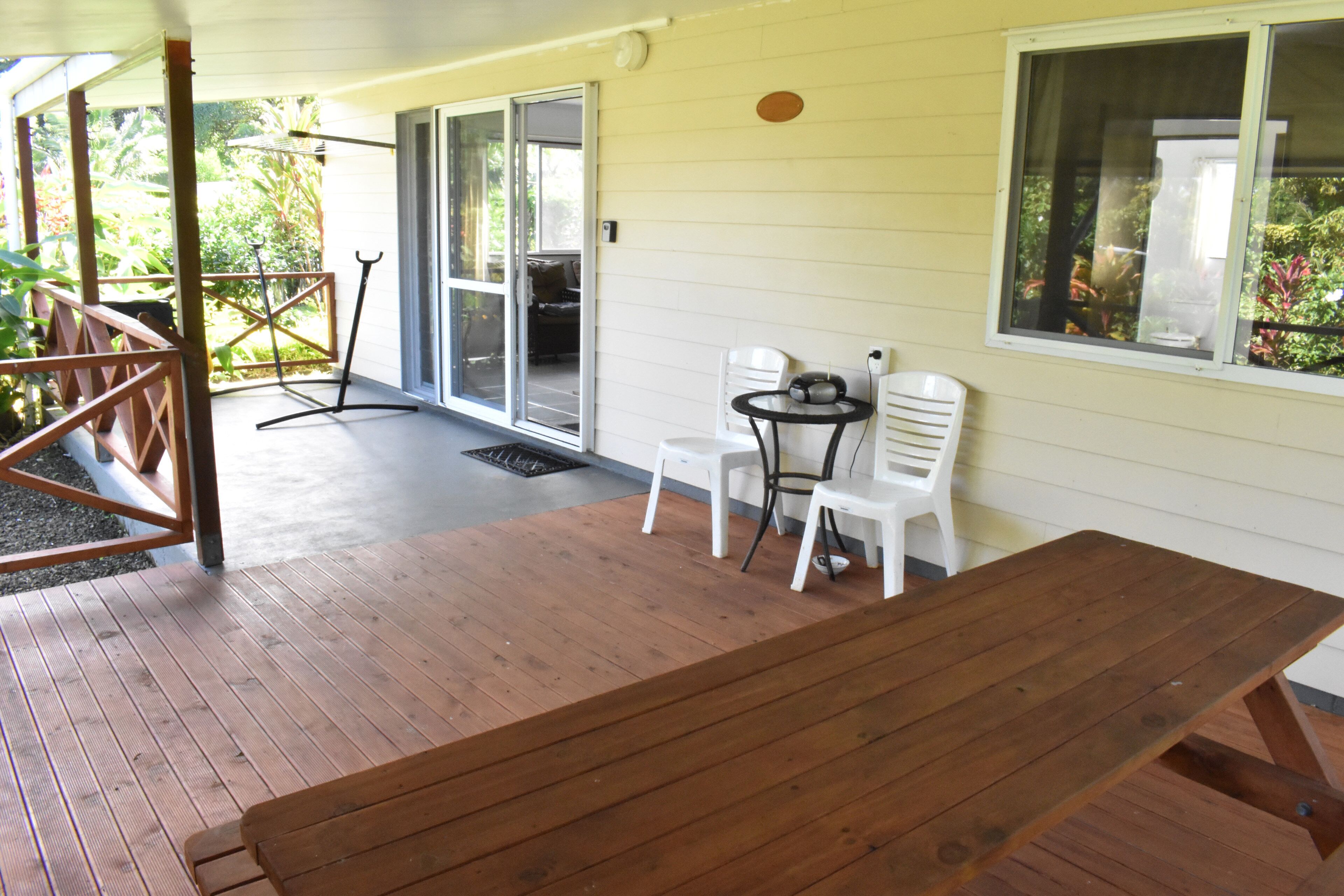 Eine Holzterrasse mit Picknicktisch und zwei weißen Stühlen, ein kleiner Tisch mit einem Radio und eine Glastür zu einem überdachten Patio.