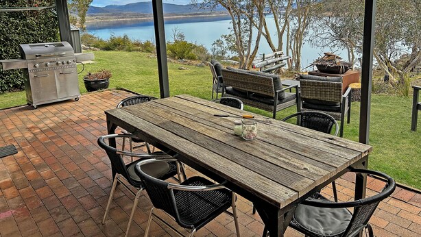 An outdoor patio with a grill, dining table, and chairs, overlooking a lake and hills.