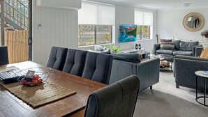 A modern living room with a wooden dining table, grey sofas, and a large TV screen.