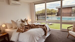 A bedroom with a large bed, a nightstand, a lamp, and a view of a swimming pool outside. - Devonlea Olive Grove
4br Home with pool on 8acres- next door to The Cups Estate (Fingal)