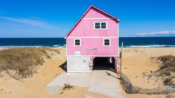 A pink beach house with a single door and a small porch, located on a sandy beach with the ocean in the background. - Right on the Beach and Dog Friendly! (Kitty Hawk)
